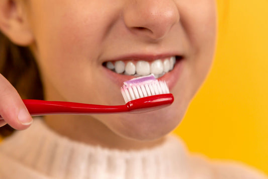 "Close-up of a person smiling with white teeth while holding a red toothbrush with toothpaste against a vibrant yellow background."