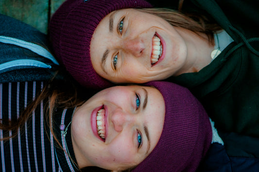 Two friends lying side by side, smiling brightly with radiant white teeth, wearing cozy purple beanies and enjoying a joyful moment together.