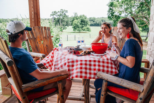 "Three friends enjoying an outdoor meal on a sunny day, smiling and sharing snacks around a table with a red and white checkered tablecloth."