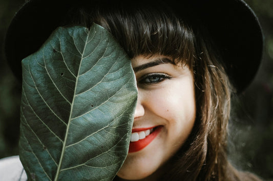 Close-up of a smiling woman with bright red lipstick, partially hiding her face behind a large green leaf, showcasing her radiant white teeth.